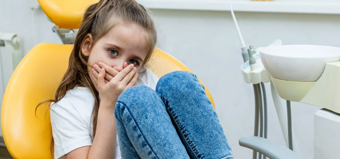 Cute little girl sitting in the dentist chair and covering her mouth in fear, being afraid of a dental treatment